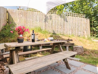 An outdoor wooden picnic table with a flower pot wine bottle glasses a hat and sunglasses near a wooden fence at Dart View Hideout in Totnes