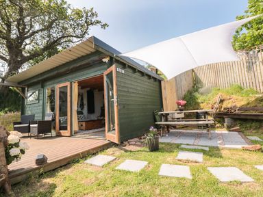 A small wooden cabin with open doors a seating area and a picnic table under a shade sail at Dart View Hideout in Totnes