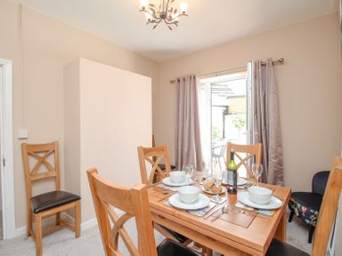 A dining room with a wooden table set for four and chairs with cushions next to curtains and glass doors at The Skipper's House in Weymouth