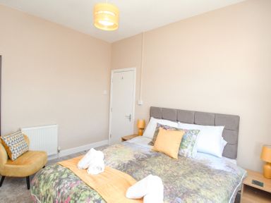 A bedroom with a bed with multiple pillows bedside tables with lamps a yellow chair and a closed door at The Skipper's House in Weymouth