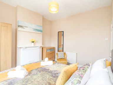 A bedroom with a bed with yellow and patterned pillows a yellow chair a wooden dresser a wardrobe a white mantel with flowers and a beach painting at The Skipper's House in Weymouth