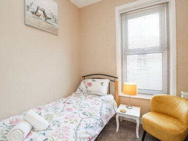 A small bedroom with a single bed with floral bedding a white side table with a lamp and a yellow chair by a window with blinds at The Skipper's House in Weymouth
