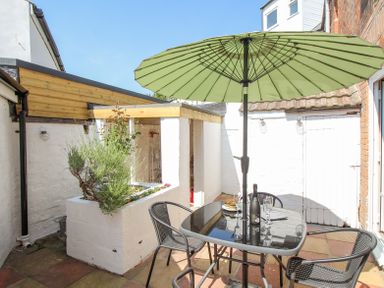A small outdoor patio with a glass table and chairs under a green umbrella and a planter box at The Skippers House in Weymouth