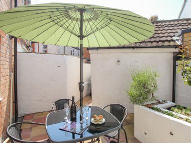A patio area with a glass table and chairs under a large green umbrella with a plant in a planter at The Skippers House in Weymouth