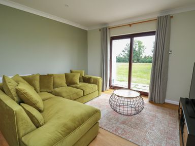 A living room with a green sectional sofa a wireframe coffee table a patterned rug and sliding glass doors with curtains at Sea View in St Clears