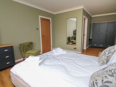A bedroom with a bed with white linens pillows folded towels a green chair wooden door black dresser and wardrobe and a wall mirror at Sea View in St Clears