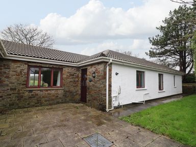 A single-story house with stone and white exterior walls and a tiled roof with a paved patio and green lawn at The Oaks in St Clears