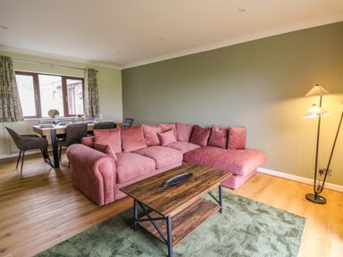 A living room with a pink sectional sofa and wooden coffee table next to a dining area with a table and chairs at The Oaks in St Clears