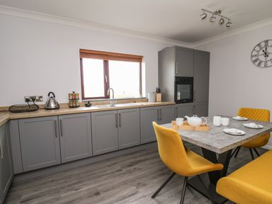 A kitchen with gray cabinets a sink under a window a built in oven and a dining table with yellow chairs at The Oaks in St Clears
