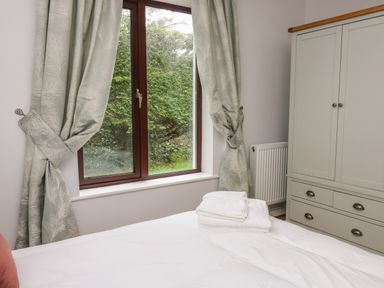 A bedroom with tied curtains on a window showing greenery outside a folded towel on a bed and a wardrobe at The Oaks in St Clears