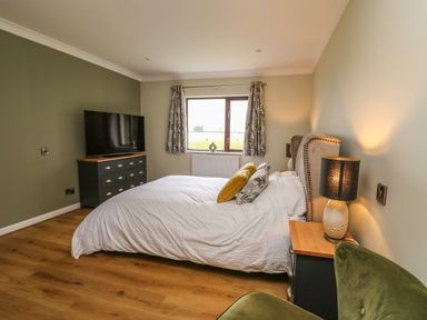 A bedroom with a bed white bedding and pillows a green armchair a window with curtains a dresser with a television and bedside tables with lamps at The Oaks in St Clears