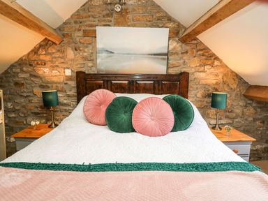A bedroom with a wooden bed frame white bedding round green and pink pillows stone walls and wooden side tables with lamps at The Bake House in Bettws