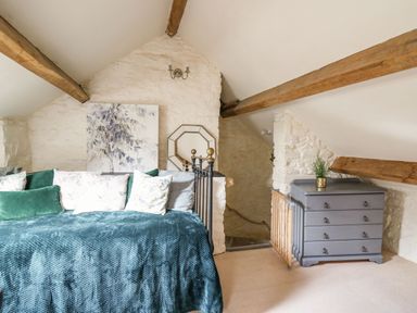 An attic bedroom with a bed covered in blue and green blankets and cushions a gray dresser with a plant and exposed wooden beams at The Bake House in Bettws