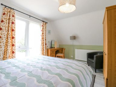 A bedroom with a bed and patterned curtains next to a window a wooden desk with a chair a wardrobe a black armchair and a floor lamp at Kingfisher in Isleham