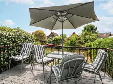 An outdoor deck with a table an umbrella and four cushioned chairs overlooking greenery and houses at Kingfisher in Isleham