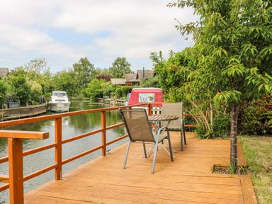 A wooden deck with two chairs and a round table by a river with boats and trees at Kingfisher Isleham