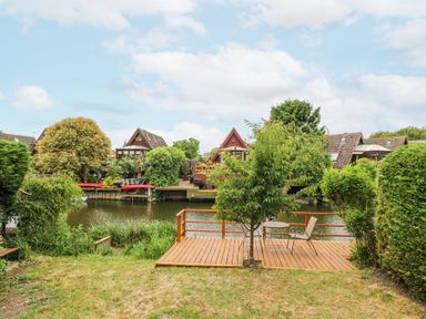 A backyard with a wooden deck overlooking a river with houses on the opposite bank at Kingfisher in Isleham