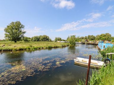 A river with boats docked along the bank and sheep grazing on grass in the field at Kingfisher in Isleham