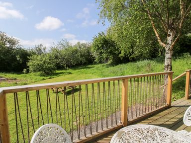 A wooden deck with white metal table and chairs overlooking a grassy yard with trees at Mucklewick Hollow in Minsterley near Bishops Castle