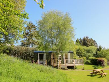 A cabin with a wooden deck surrounded by trees and grass with a picnic table in the foreground at Mucklewick Hollow in Minsterley near Bishops Castle