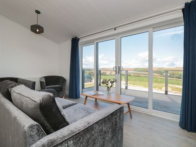 A living room with a gray sofa and chair a wooden coffee table with flowers and a magazine and large glass doors overlooking fields at Bluebell Lodge in Norton near Dartmouth