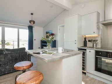 A kitchen area with a countertop, two wooden stools, a vase with flowers, and a gray couch near large glass doors at Bluebell Lodge in Norton near Dartmouth