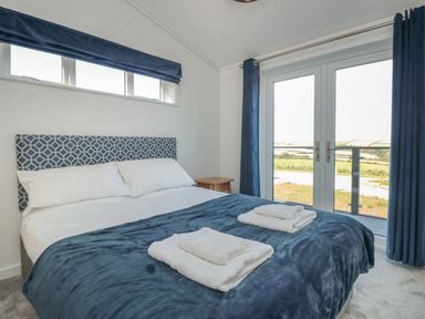 A bedroom with a double bed with white linens and folded towels two windows and blue curtains at Bluebell Lodge in Norton near Dartmouth