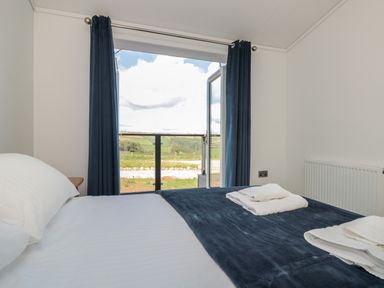 A bedroom with a bed covered in white and dark blue linens towels on the bed and open glass doors with blue curtains at Bluebell Lodge Norton near Dartmouth