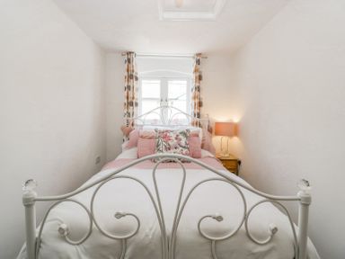 A bedroom with a white metal bed frame pink bedding a wooden nightstand with a lamp and window with patterned curtains at 4 Wallflower Row Mordiford near Hereford