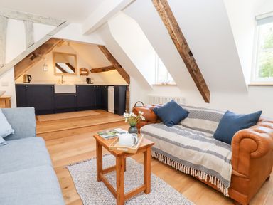 A living area with two sofas a wooden coffee table and a kitchen with dark cabinets under a sloped ceiling with wooden beams at Spring Cottage in Stroud