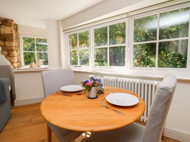 A dining area with a round wooden table set for two near windows showing greenery outside at Spring Cottage in Stroud