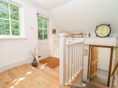 A landing area with wooden flooring a white door window a vase with branches white railing stairs and a clock on a shelf at Spring Cottage in Stroud