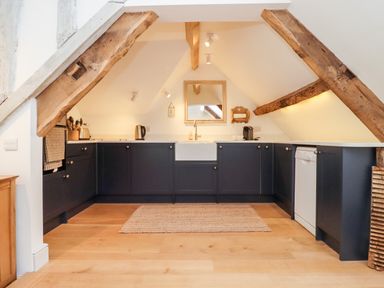 A kitchen with dark lower cabinets a farmhouse sink wooden beams on the ceiling and a rug on the floor at Spring Cottage in Stroud