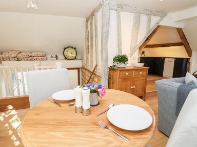 A dining area with a round wooden table set for two with white plates salt and pepper shakers and flowers a wooden cabinet with a plant and bowl and a kitchen with black cabinets in the background at Spring Cottage in Stroud