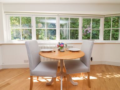 A dining area with a round wooden table and two gray upholstered chairs near large windows with a view of greenery at Spring Cottage in Stroud