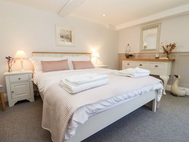 A bedroom with a double bed having white and pink bedding two bedside tables with lamps and flowers a dresser with a mirror and decorations at Spring Cottage in Stroud