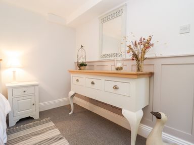 A bedroom corner with a bedside table and lamp next to a wooden dresser with a mirror and decorative items at Spring Cottage in Stroud