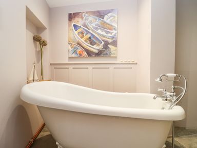 A bathroom with a white freestanding bathtub a painting of boats on the wall and decorative items in a niche at Spring Cottage in Stroud