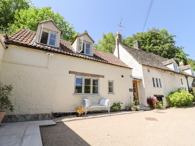 Exterior view of a cream-colored stone cottage with two dormer windows and a bench under the main window at Spring Cottage in Stroud