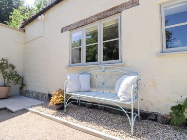 A metal bench with cushions outside a stone house with windows and potted plants at Spring Cottage in Stroud