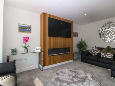 A living room with a mounted television, a wooden panel fireplace, black sofas, a glass console table with a pink flower, and a round rug at Hedd in Amlwch
