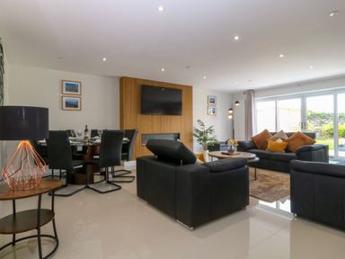 A living room with black sofas orange cushions a round wooden table a TV on a wooden panel and a dining table with black chairs at Hedd in Amlwch
