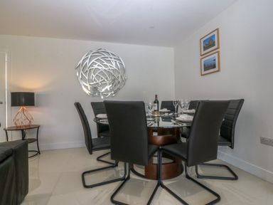 A dining room with a round glass table surrounded by six black chairs and a lamp on a side table at Hedd in Amlwch