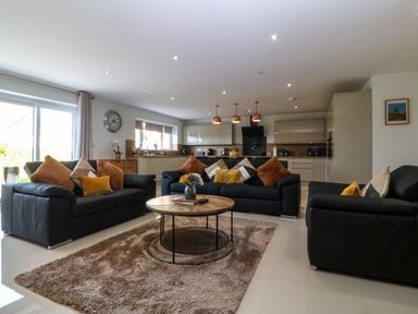 A living room with black sofas and brown cushions around a wooden coffee table and a kitchen area in the background at Hedd in Amlwch