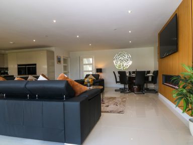An open living room with black leather sofas and orange pillows a dining area with black chairs a wall-mounted TV and fireplace and a plant on the floor at Hedd in Amlwch