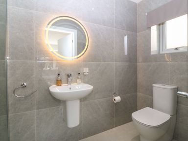 A bathroom with a wall-mounted sink, a round illuminated mirror, and a toilet at Hedd in Amlwch