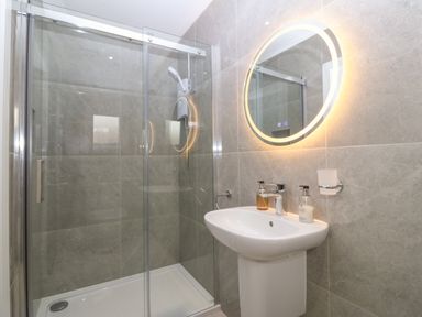 A bathroom with a glass shower enclosure a pedestal sink and a round illuminated mirror at Hedd in Amlwch