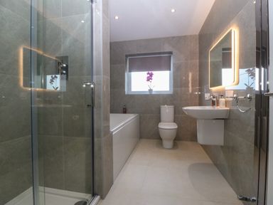 A bathroom with a shower enclosure bathtub toilet sink and illuminated mirror with a window showing a flower at Hedd in Amlwch