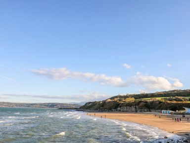 A sandy beach with waves and people walking along the shoreline with cliffs and hills in the background at Hedd in Amlwch