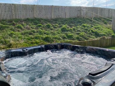 An outdoor hot tub with bubbling water next to a grassy hill and wooden fence at Galwad y Mor in Broad Haven